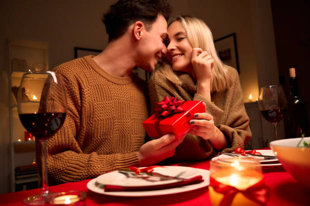 glückliches junges paar feiert jubiläum oder valentinstag mit romantischem abendessen zu hause tisch. liebender mann gibt rote geschenk-box umarmung geliebte frau machen gegenwart überraschung auf datum in kerzenlicht. - food and drink holidays and celebrations people valentines day stock-fotos und bilder