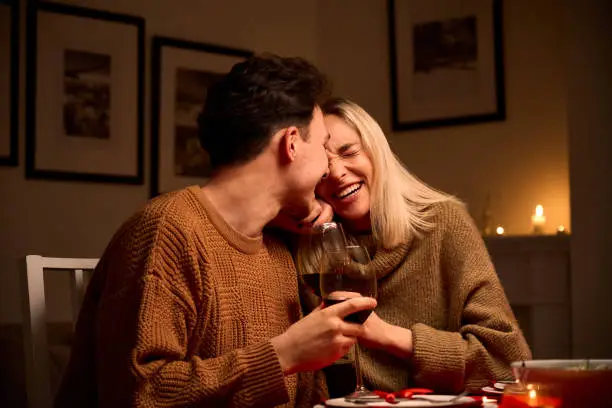 Photo of Happy young couple in love hugging, laughing, drinking wine, enjoying talking, having fun together celebrating Valentines day dining at home, having romantic dinner date with candles sitting at table.
