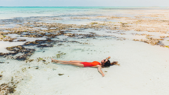 Young woman with long hair in red swimsuit and sunglasses lying in the pure turquoise Indian Ocean on Zanzibar island, Tanzania during bright summer day