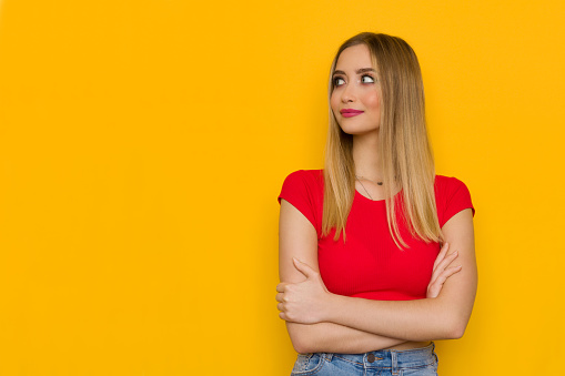 Young blond woman in red top is standing relaxed with arms crossed and looking away. Front view. Waist up studio shot on yellow background.