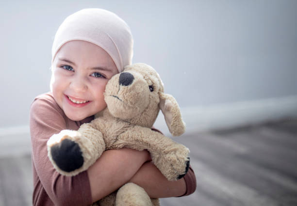 My favourite teddy bear! A young girl who has leukaemia is sitting on the floor in her room. She is wearing a headscarf due to her hair loss and is holding onto her favourite teddy bear. She is smiling widely at the camera. cancer illness stock pictures, royalty-free photos & images