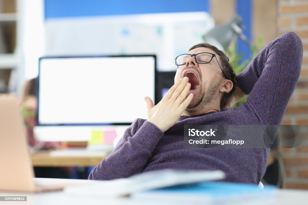 Young man siting at table in office and yawning Young man siting at table in office and yawning. Procrastination concept Yawning Stock Photo