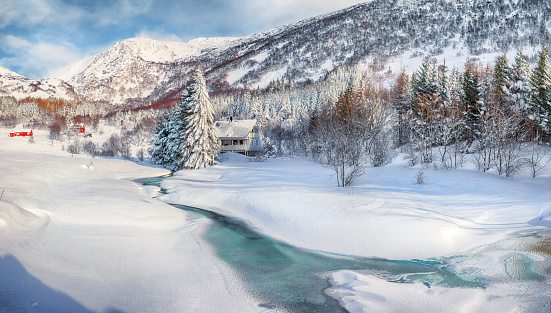Awesome winter scenery with frozen river with wooden houses and snow covered pine trees near Valberg village at Lofotens.  Location: Valberg, Vestvagoy, Lofotens, Norway