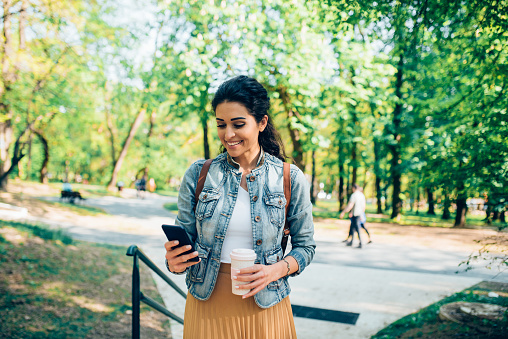 Portrait of a beautiful young woman walking in the park, drinking coffee and using a smartphone.