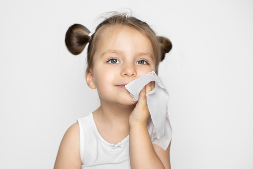 Little beautiful Caucasian blond girl with ponytails, wipes her face and cheek with a wet napkin, looking up and smiling. Studio shot on isolated white background with copy space.