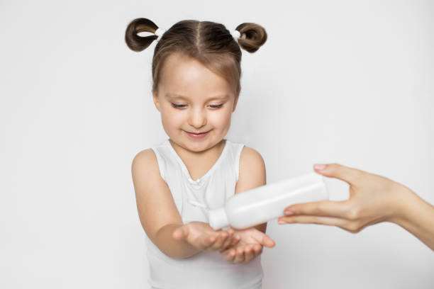 istock A little girl in white tank top and with two ponytails, standing on white background while hand of her mom pours a sanitizer on her hands. Sanitary precautions - hand disinfection. 1297602003