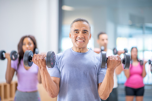 A male senior of Hispanic ethnicity is participating at a group fitness class. He is doing using a dumbbell to exercise.