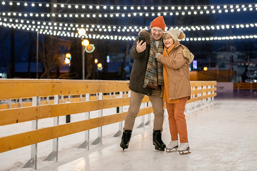 Happy mature couple making photo on mobile phone during his ride on ice rink in the park