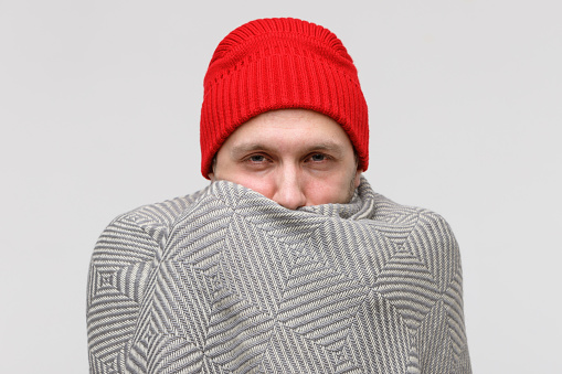 Sick man in red beanie wrapped in warm beige knitted plaid, suffering from cold, looking at camera, isolated on gray background. Eyes are red and sore from the disease. Flu season.