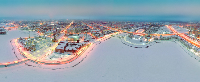 Buildings decorated with turrets and domes and Orthodox churches located on the central embankment in the city center, surrounded by residential areas. The aerial panoramic view of cityscape with night illumination, snowy winter, evening. Yoshkar-Ola, Volga region, Russia