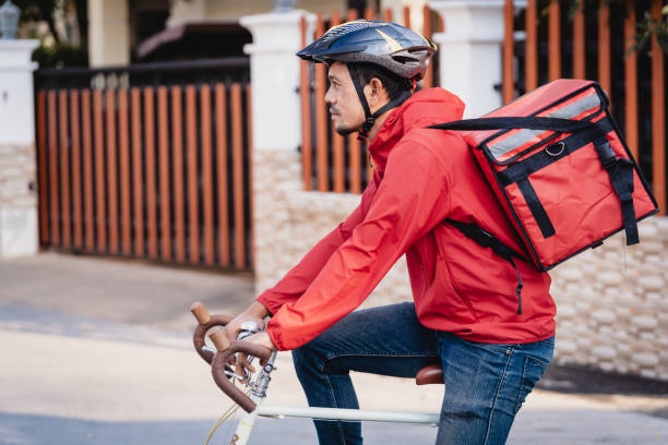 istock Courier in red uniform with a delivery box on back riding a bicycle and looking on the cellphone to check the address to deliver food to the customer. Courier on a bicycle delivering food in the city. 1297620685