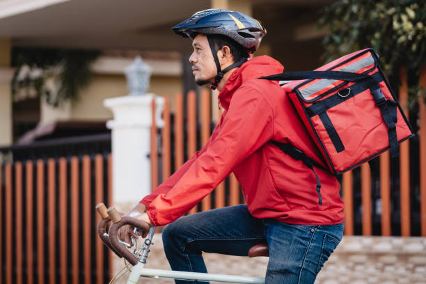 istock Courier in red uniform with a delivery box on back riding a bicycle and looking on the cellphone to check the address to deliver food to the customer. Courier on a bicycle delivering food in the city. 1297620787