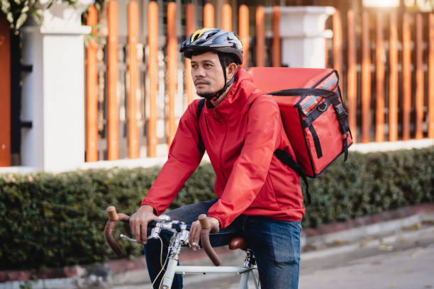 istock Courier in red uniform with a delivery box on back riding a bicycle and looking on the cellphone to check the address to deliver food to the customer. Courier on a bicycle delivering food in the city. 1297620875