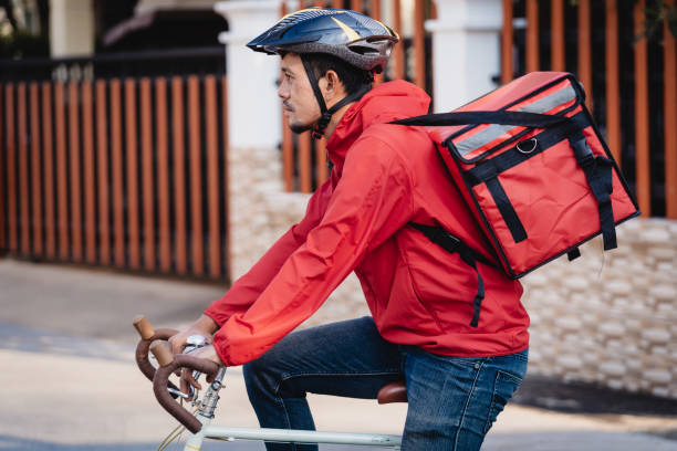 istock Courier in red uniform with a delivery box on back riding a bicycle and looking on the cellphone to check the address to deliver food to the customer. Courier on a bicycle delivering food in the city. 1297620878