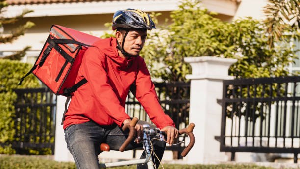 istock Courier in red uniform with a delivery box on back riding a bicycle and looking on the cellphone to check the address to deliver food to the customer. Courier on a bicycle delivering food in the city. 1297620957