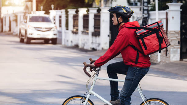 istock Courier in red uniform with a delivery box on back riding a bicycle and looking on the cellphone to check the address to deliver food to the customer. Courier on a bicycle delivering food in the city. 1297621732