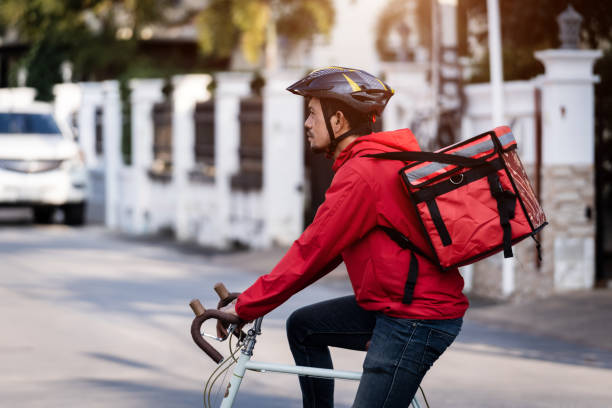 istock Courier in red uniform with a delivery box on back riding a bicycle and looking on the cellphone to check the address to deliver food to the customer. Courier on a bicycle delivering food in the city. 1297621883