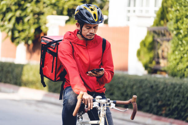 istock Courier in red uniform with a delivery box on back riding a bicycle and looking on the cellphone to check the address to deliver food to the customer. Courier on a bicycle delivering food in the city. 1297621942