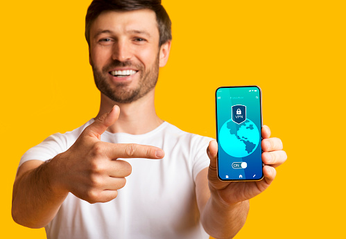 Protection Of Information And Cyber Security Online. Smiling young man holding, showing and pointing at cellphone with virtual private network application connected to local network over the internet