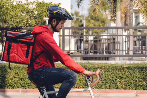 istock Courier in red uniform with a delivery box on back riding a bicycle and looking on the cellphone to check the address to deliver food to the customer. Courier on a bicycle delivering food in the city. 1297622795