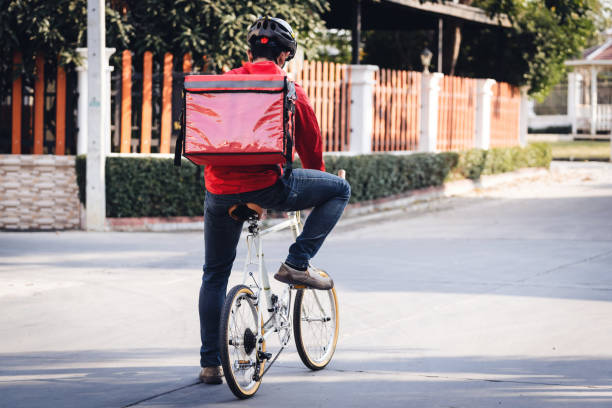 istock Courier in red uniform with a delivery box on back riding a bicycle and looking on the cellphone to check the address to deliver food to the customer. Courier on a bicycle delivering food in the city. 1297622949