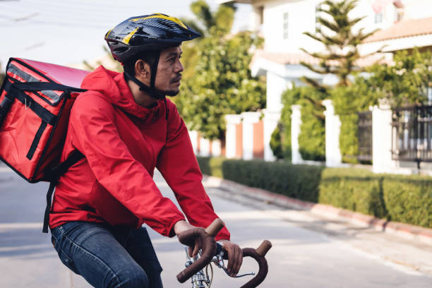 istock Courier in red uniform with a delivery box on back riding a bicycle and looking on the cellphone to check the address to deliver food to the customer. Courier on a bicycle delivering food in the city. 1297622950