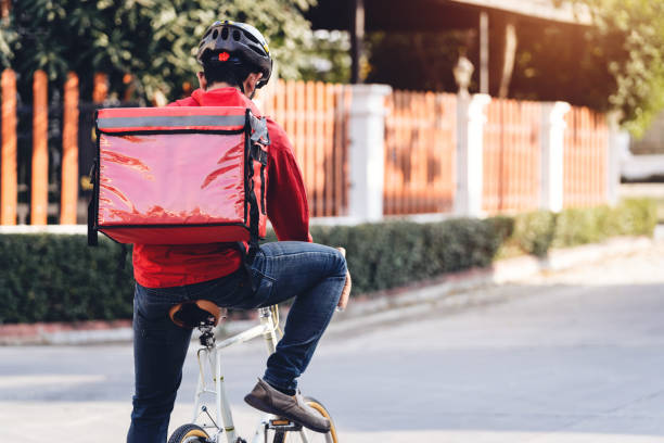 istock Courier in red uniform with a delivery box on back riding a bicycle and looking on the cellphone to check the address to deliver food to the customer. Courier on a bicycle delivering food in the city. 1297622955