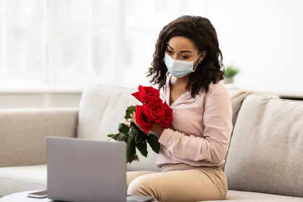 Photo of Black woman in medical mask having online date, holding roses