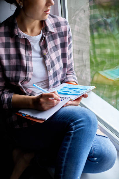 istock Woman sitting at the window, writing in notebook plan, to do list after quarantine end, coronovirus ending 1297623192