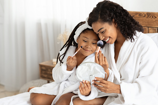 African mother and her little daughter in bathrobes making makeup on bed, having fun at home, loving black mom teaching her child beauty tricks, happy family enjoying spending time together, closeup