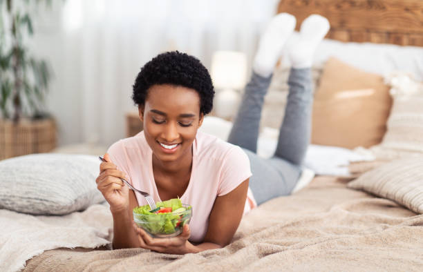 istock Attractive black lady eating vegetable salad while lying on comfortable bed at home, copy space 1297624717