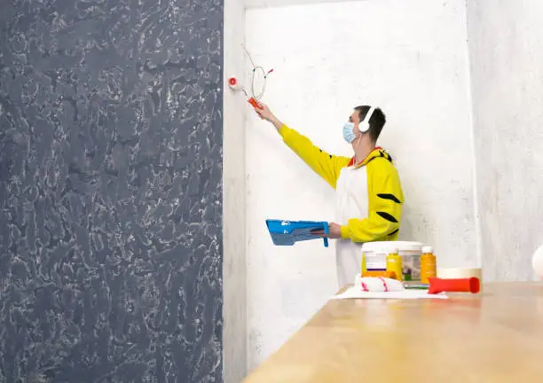 Painter man paint the wall in the room. Man in a protective mask in quarantine makes repairs at home