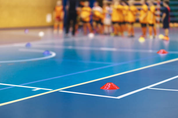istock Indoor Soccer Class. Children on Physical Education Class with Teacher Blurred Background. Soccer Training Cone Marker on Wooden Field Court 1297629720