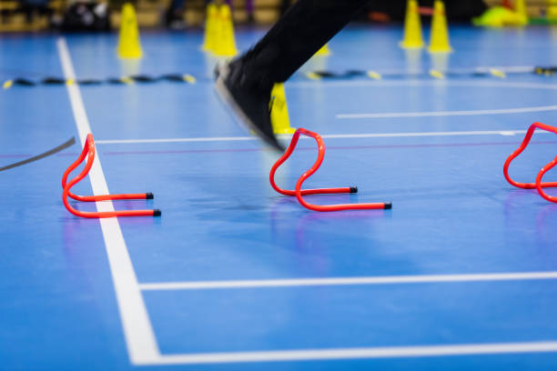 istock Athlete jumping over red training hurdles on indoor practice session. Futsal training equipment in the background. Soccer hurdles, cones and ladder on training court 1297629721