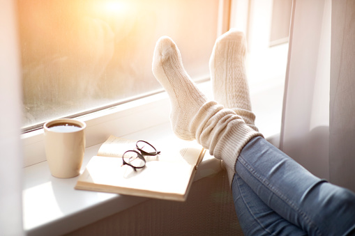 Cropped view of woman putting her feet in warm comfy socks on windowsill with book and coffee, closeup. Unrecognizable lady spending cold day at home, enjoying cozy atmosphere