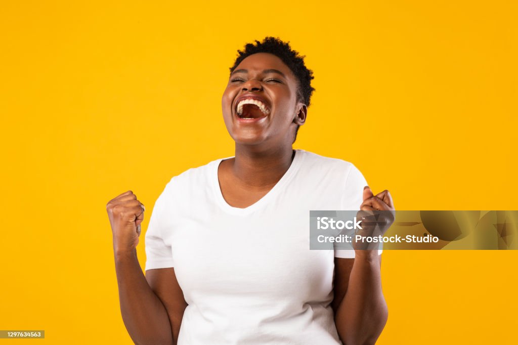 Joyful Black Woman Shouting Shaking Fists Posing Over Yellow Background Joy Of Victory. Joyful Oversized Black Woman Shouting Gesturing Yes And Shaking Fists Posing Standing Over Yellow Background, Studio Shot. Excitement And Joy, Success Celebration. Emotional Portrait. Women Stock Photo