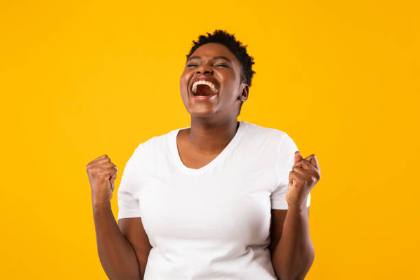 joyful black woman shouting shaking fists posing over yellow background - women standing fist success imagens e fotografias de stock