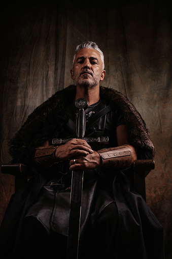 Studio shot a strong fantasy knight with sword with male dramatic gaze looking down from above sitting on a simple wooden throne. For historical novel concepts. Creative dramatic color retouching to underline the ancient medieval time. Vignetting and added noise. Part of a series.