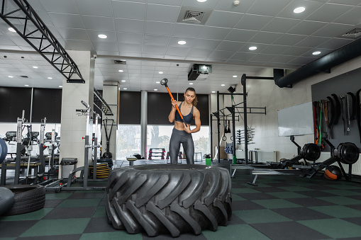 Cross, functional training. Young fit woman hits the wheel with a heavy hammer in a modern cross gym