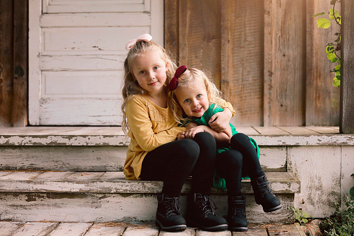 Two sisters, ages five and three, smile at the camera as they sit on a porch. Shot with a Canon 5D Mark iv.