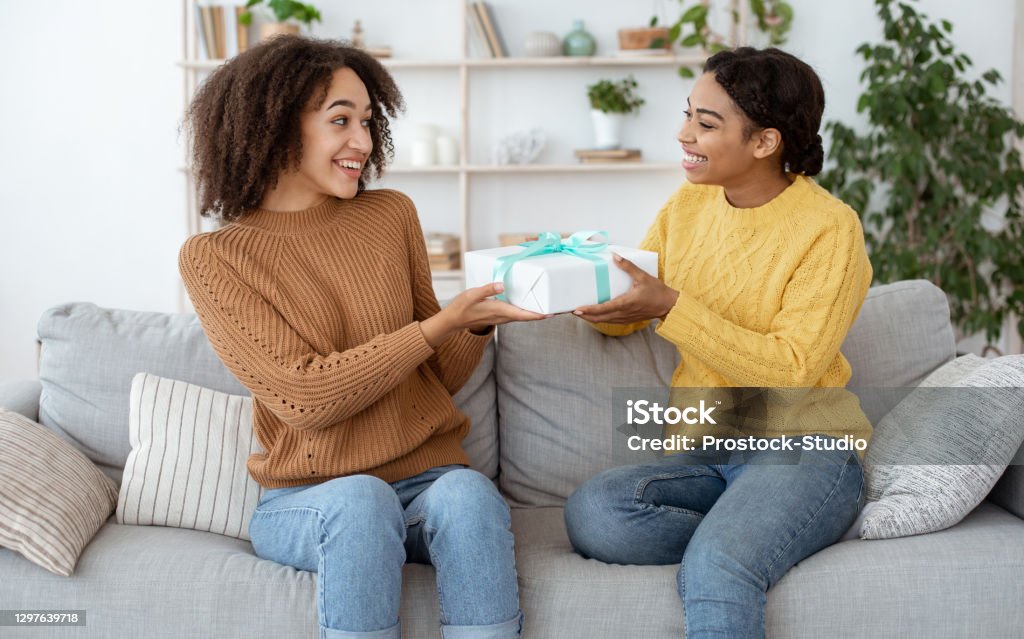 Two ladies friends making surprise birthday present Two ladies friends making surprise birthday present. Happy millennial african american female congratulates, gives gift to embarrassed mixed race sister, sits on sofa in interior of living room Gift Stock Photo
