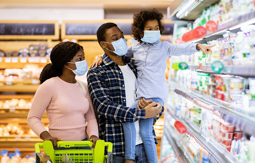 Family shopping during coronavirus pandemic. African American family with child wearing face masks, purchasing food at supermarket, panorama. Black parents with their kid buying products at mall