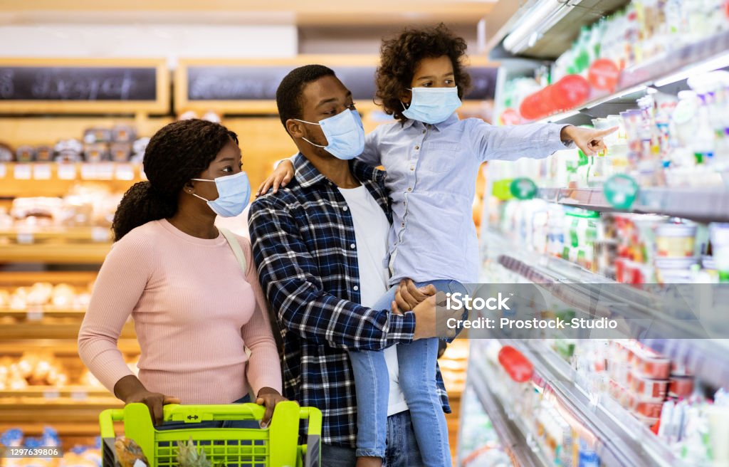 Family shopping during coronavirus pandemic. Black family with child wearing face masks, purchasing food at supermarket Family shopping during coronavirus pandemic. African American family with child wearing face masks, purchasing food at supermarket, panorama. Black parents with their kid buying products at mall Protective Face Mask Stock Photo