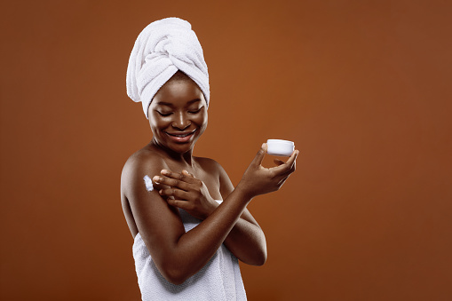 Skincare Products. Beautiful African American Lady With Towel On Head Applying Moisturizing Body Cream After Shower, Smiling Black Woman Nourishing Skin After Bath, Standing Over Brown Background