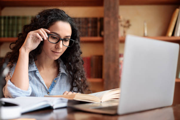 Education And Pastime Concept. Portrait of young woman reading paper book, touching eyeglasses, sitting at the desk at home office or library, enjoying story, stuying and learning with laptop on table