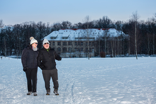 Couple walks in an open field during a winter afternoon, they looks happy and joyful. They are in an open forest in Germany and a private building outside the park serves as a background.