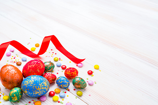 Holidays: hand painted multi colored Easter eggs arranged at the bottom left corner of a white striped table leaving useful copy space for text and/or logo. Some candies, sugar sprinkles and a red ribbon complete the composition. High resolution 42Mp studio digital capture taken with Sony A7rII and Sony FE 90mm f2.8 macro G OSS lens