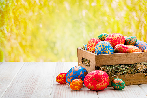 Holidays: Hand painted multi colored Easter eggs in a crate shot outdoors on striped white table against defocused yellow lush foliage. The composition is at the right of an horizontal frame leaving useful copy space for text and/or logo at the left. High resolution 42Mp outdoors digital capture taken with Sony A7rII and Sony FE 90mm f2.8 macro G OSS lens