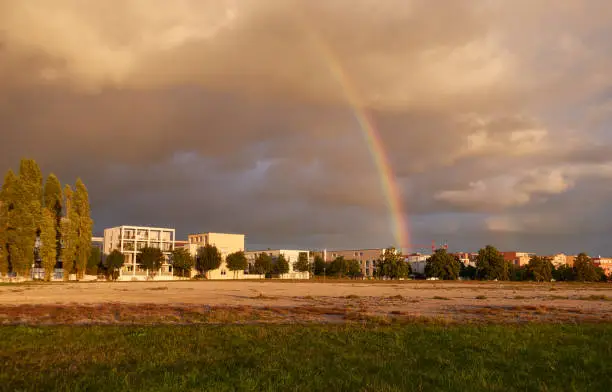 Rainbow against a gray cloudy evening sky, housing estate with square-shaped residential buildings, terrain with pebbles and green meadow in the foreground, tall trees on the edge. Germany, Ostfildern