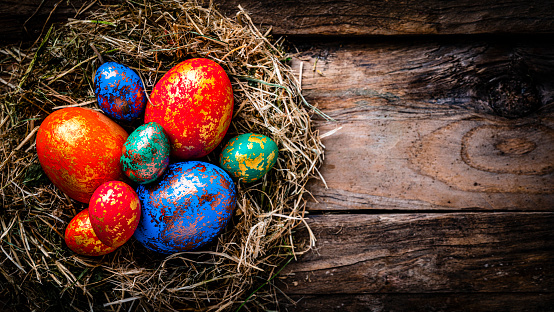 Holidays: Hand painted multi colored Easter eggs in a straw nest shot from above on rustic wooden table. The composition is at the left of an horizontal frame leaving useful copy space for text and/or logo at the right. High resolution 42Mp studio digital capture taken with Sony A7rII and Sony FE 90mm f2.8 macro G OSS lens